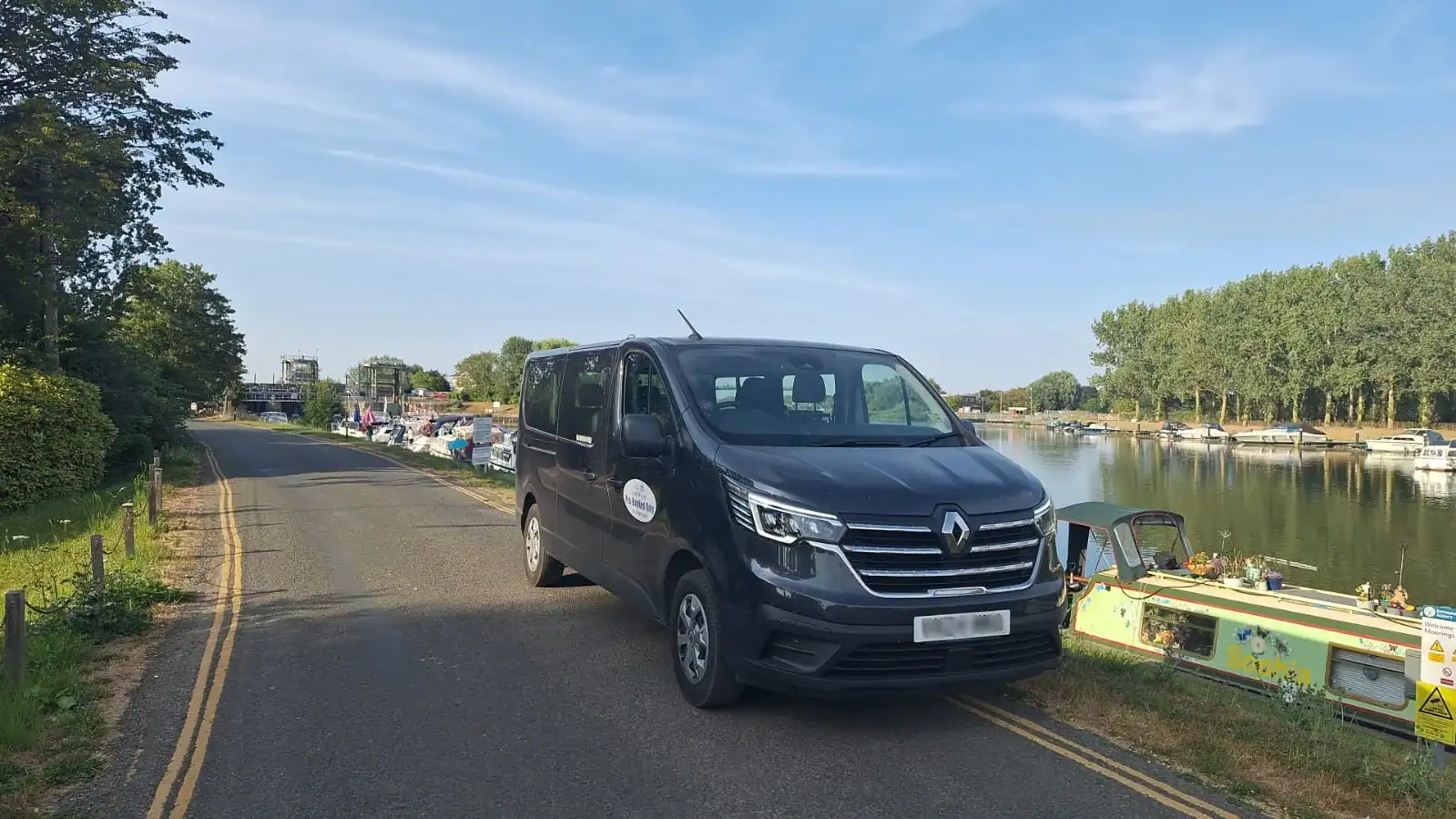 Minibus hire Downham Market – G and M Taxis dark grey Renault minibus parked beside a riverside road with boats in the Norfolk sunshine.
